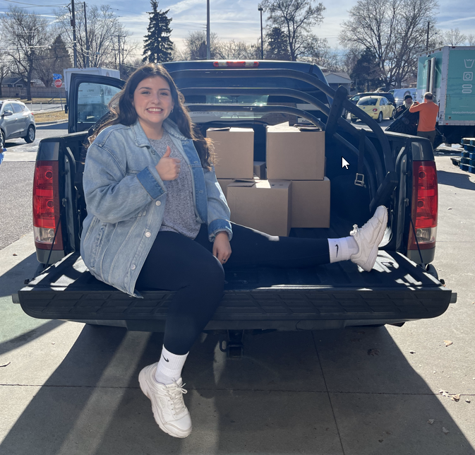 An image of a woman sitting in the back of a pick up truck with 7 boxes of donation items